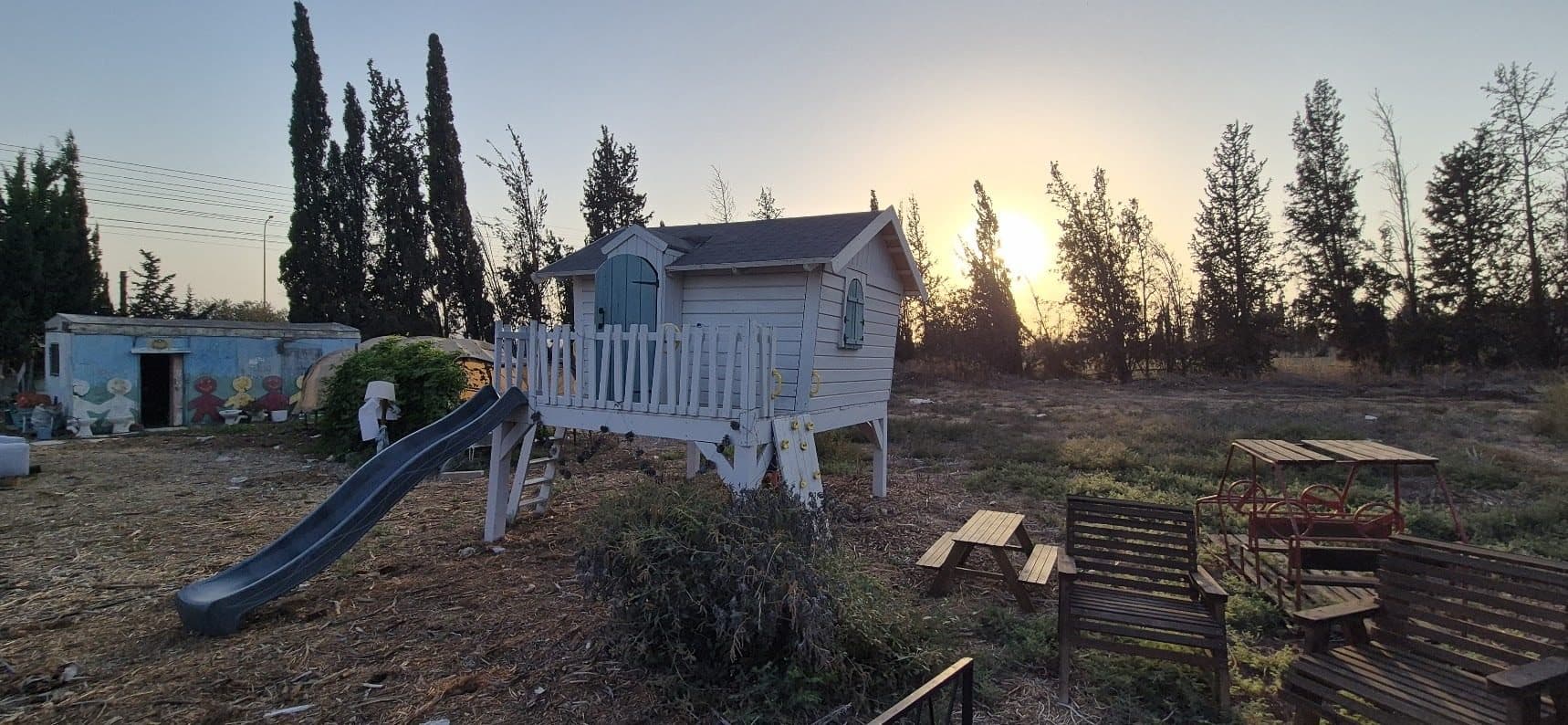 שקיעה בגינה הקהילתית בקיבוץ גניגר: בית העץ הלבן, ספסלי עץ ושורת ברושים