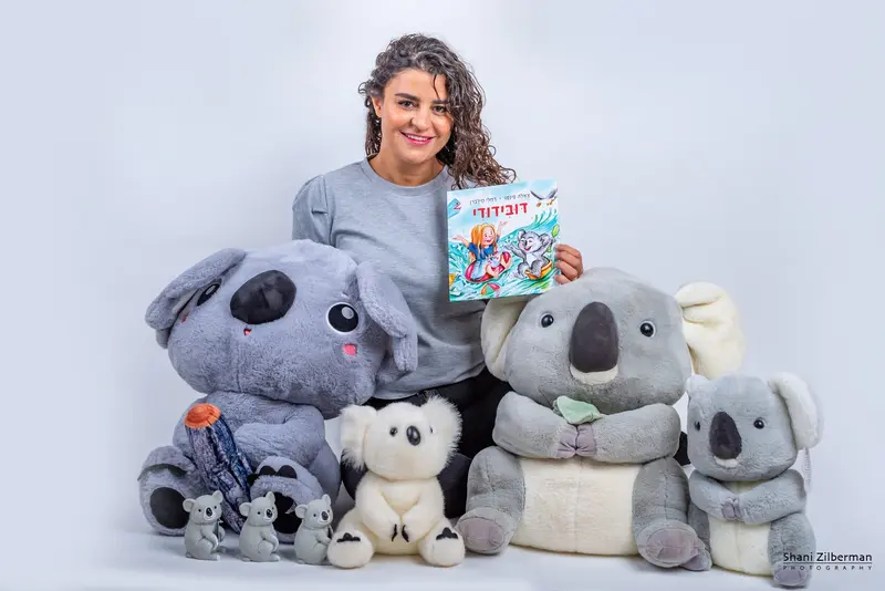 הסופרת מחזיקה את הספר דובידודי, מוקפת בבובות קואלה